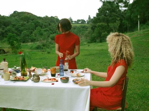As duas mulheres das fotos anteriores aparecem juntas em um cenário campestre. Ambas vestem roupas vermelhas vibrantes e estão em uma mesa de jantar montada ao ar livre sobre um gramado verde. Uma delas está sentada de perfil, enquanto a outra está de pé ao lado da mesa, manuseando uma garrafa. A mesa tem uma toalha branca e está servida com diversos pratos e copos. Ao fundo, vê-se uma paisagem de colinas arborizadas sob um céu claro, transmitindo uma sensação de tranquilidade e natureza.