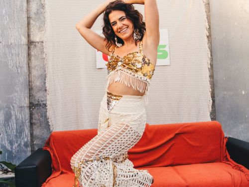 Fotografia colorida da cantora Mariana Aydar posando sorridente à frente de um sofá com manta laranja. Ela usa um conjunto artesanal de duas peças em tons de branco e dourado: um top bordado com franjas e uma calça de trama vazada (crochê). Ela está com os braços erguidos sobre a cabeça e uma perna levemente flexionada. O cenário é urbano e ao ar livre, com prédios ao fundo sob a luz do dia.