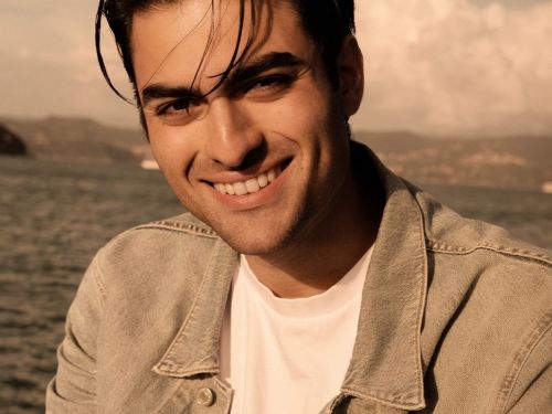 Retrato em plano médio do cantor Matteo Bocelli ao ar livre. Ele aparece sorrindo, com o rosto levemente inclinado, vestindo uma camiseta branca sob uma jaqueta jeans de lavagem clara. O fundo possui um tom sépia quente, mostrando uma paisagem costeira com o mar, montanhas distantes e um céu com nuvens suaves. A iluminação é natural, destacando seus traços e o cabelo escuro levemente desalinhado.