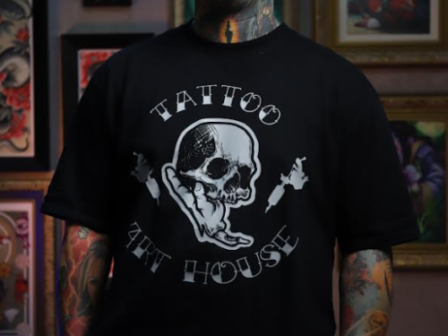 Um homem jovem e sorridente posa em um estúdio de tatuagem. Ele possui diversas tatuagens no rosto, pescoço e braços, além de alargadores pretos nas orelhas. Veste uma camiseta preta com a estampa de uma caveira sendo segurada por uma mão e o texto "Tattoo Art House". Ao fundo, a parede está decorada com vários quadros de artes de tatuagem e um relógio neon circular.