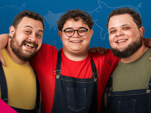 Três homens jovens e sorridentes aparecem abraçados do peito para cima, transmitindo alegria e união. Todos vestem macacões jeans sobre camisetas coloridas: o da esquerda veste amarelo, o do centro (que usa óculos redondos) veste vermelho, e o da direita veste verde-oliva. O fundo é azul com silhuetas sutis de porquinhos e um detalhe geométrico rosa no canto inferior.