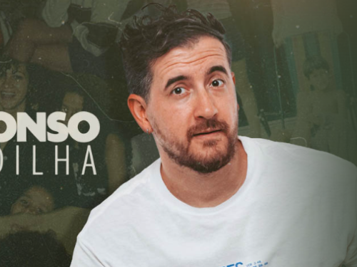 A imagem mostra o comediante brasileiro Afonso Padilha em primeiro plano, centralizado à direita. Ele é um homem branco de cabelos castanhos curtos e levemente bagunçados, barba aparada e usa uma camiseta branca simples. Ele olha para a câmera com uma expressão de dúvida ou questionamento.