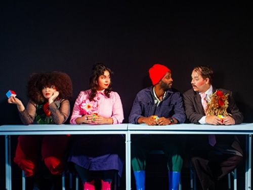 Quatro atores sentados atrás de uma mesa, imagem com fundo preto. Duas mulheres à esquerda e dois homens à direita. As mulheres vestem blusa longa e saia, os homens usam ternos e acessórios coloridos. 