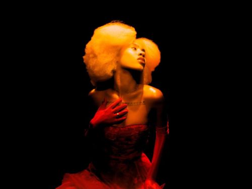 Mulher com cabelo volumoso e claro posa em fundo totalmente preto. Iluminada por luz quente, veste um vestido vermelho e luvas longas, com uma das mãos sobre o peito e o rosto inclinado para cima, criando uma cena dramática e teatral.