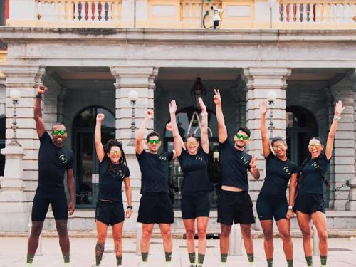 Sete atletas corredores em frente à Praça Rui Barbosa, há três homens e quatro mulheres. Eles vestem blusa e bermuda pretas e erguem um braço em comemoração.