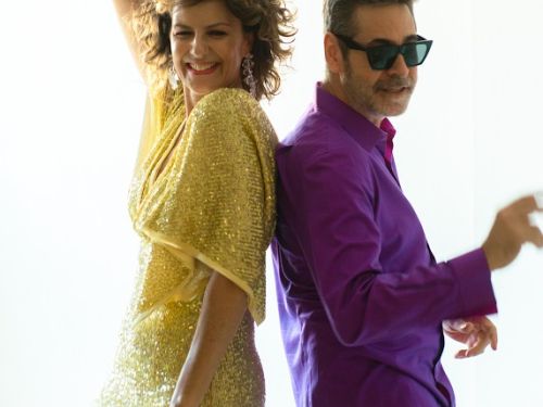 Mulher e homem apoiados de costas. A mulher (à esquerda) usa vestido amarelo e o homem (à esquerda) usa blusa social longa roxa, calça clara e óculos escuros. Ambos gesticulam passos de dança.