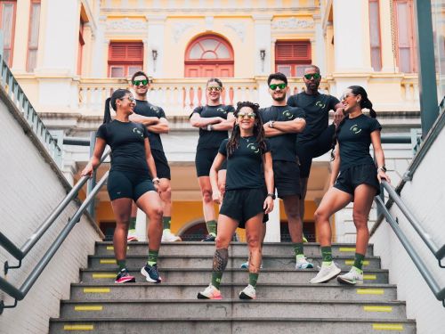 Sete atletas corredores na escadaria da Praça Rui Barbosa, há três homens e quatro mulheres. Eles vestem blusa e bermuda pretas e sorriem de braços cruzados.