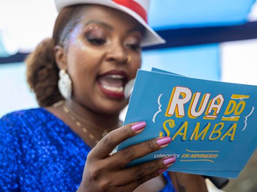 Mulher com roupa azul brilhante e chapéu branco fala ao microfone enquanto lê um cartão com o título “Rua do Samba – alegria em movimento”. Ela usa maquiagem com brilho e brincos grandes.