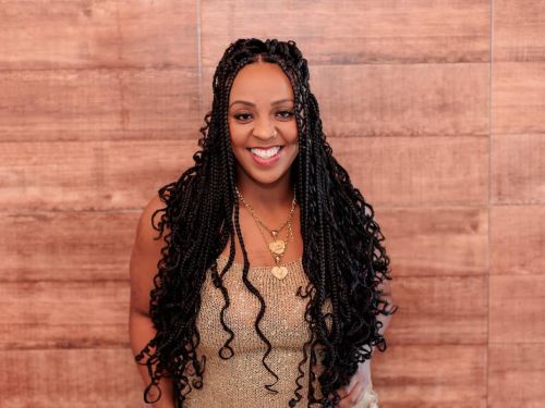 Artista Thata Ferreira. Mulher preta sorrindo, veste blusa marrom e acessório dourado no pescoço. Foto sob fundo marrom.