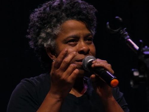 Escritora Nívea Sabino. Mulher preta segurando um microfone enquanto gesticula com a outra mão.