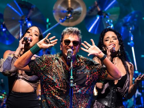 Cantor ao centro acompanhado de duas mulheres, uma em cada lado, reproduzindo vocal de apoio. Eles vestem roupas escuras com estampa, há instrumentos musicais ao fundo.