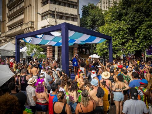 Multidão reunida em um evento de rua ao ar livre. No centro, um pequeno palco coberto por estrutura azul e branca recebe músicos, enquanto o público, com roupas leves e coloridas, ocupa a praça ao redor. Prédios e árvores aparecem ao fundo.