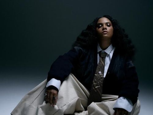 Artista DJ Kingdom. Mulher preta sentada no chão, veste terno escuro e calça clara de modelagem larga, expressão facial séria. Foto sob fundo escuro. 