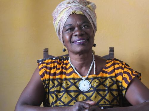 Professora Rosália Diogo. Mulher preta sentada em cadeira de madeira, ela veste blusa de manga curta com cores amarela e preta, acessórios no pescoço, orelha e turbante na cabeça.