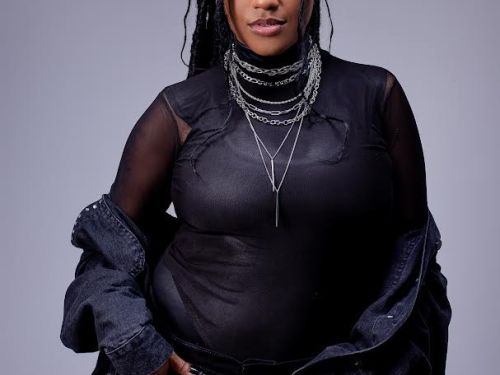 Artista Raquel Moreira. Mulher preta em pé, veste roupas pretas e acessórios pratas. Foto sob fundo cinza.