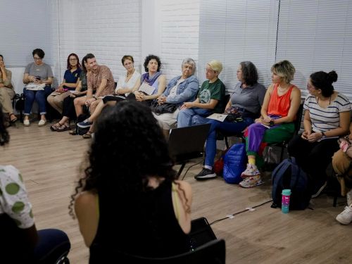 Pessoas sentadas em cadeiras em formato de roda de conversa. Ambiente fechado com paredes branca e cinza e chão marrom.