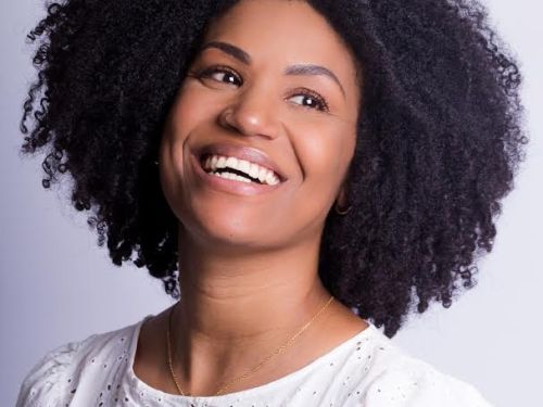 Artista Vavá Fernandes. Mulher preta sorrindo, veste blusa branca. Foto colorida sob fundo claro.