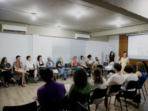 Pessoas sentadas em cadeiras em formato de roda de conversa. Ambiente fechado com paredes branca e cinza e chão marrom.