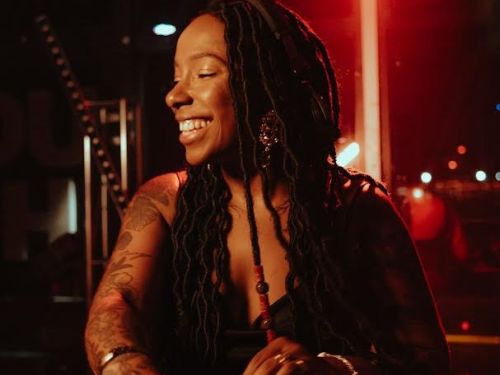 Artista DJ Camis. Mulher preta tocando em uma mesa operadora de DJ. Veste blusa escura e sorri com os olhos fechados. Foto sob fundo escuro com luzes vermelhas.