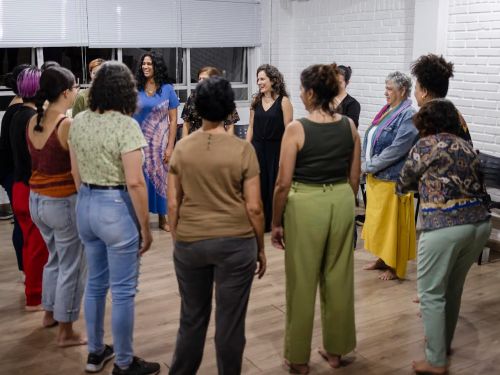 Pessoas em pé em formato de roda. Ambiente fechado com paredes branca e cinza, janelas de vidro e chão marrom.