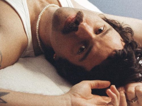 Artista Rafa Castro deitado com os braços próximos à cabeça enquanto encara a câmera com semblante sério. Veste camisa regata branca.