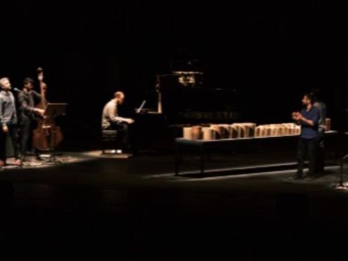 Cinco artistas no palco tocando instrumentos. Palco escuro com iluminação concetrada nos artistas.