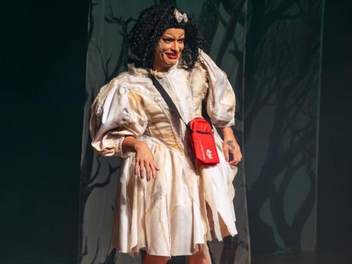 Suzy Brasil usando vestido branco com detalhes em amarelo, bolsa vermelha sobre os ombros e laço prata na cabeça. Imagem com fundo escuro.