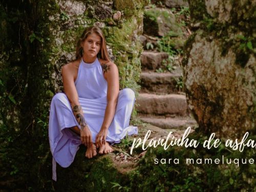 Artista Sara Mameluque sentada em um chão de pedra coberto por vegetação rasteira. Ela usa vestido lilás e os pés descalõ, encara a câmera com semblante sério.