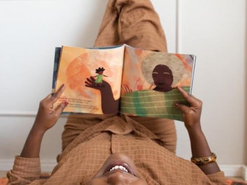 Artista Éle Fernandes (mulher preta) deitada no chão com os pés apoiados na parede branca. Ela sorri para a câmera enquanto segura o exemplar do livro "Dinari"