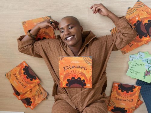 Artista Éle Fernandes (mulher preta) deitada no chão com os pés apoiados na parede branca. Ela sorri para a câmera e está rodeada de exemplares do livro "Dinari"