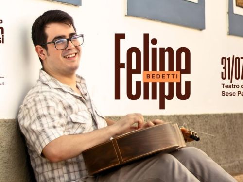 Artista Felipe Bedetti. Homem branco sentado no chão, sorrindo e com o violão sobre as penas. Veste blusa de botão flanelada e calça cinza. Costas apoiadas na parede externa de uma casa.