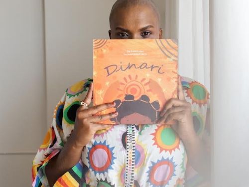 Artista Éle Fernandes (mulher preta) segurando um exemplar do livro "Dinari" em frente ao rosto. Ela veste uma blusa de estampa colorida, imagem com fundo claro.