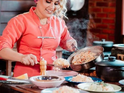 Cozinheira servindo comida em um prato enquanto segura a panela com uma das mãos. Ela veste dolma rosa, usa um pano na cabeça e está sorrindo. Ao seu redor, há panelas e pratos com comidas.
