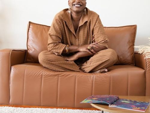 Arista Éle Fernandes (mulher preta) sentada de pernas cruzadas em um sofá marrom, ela sorri para a câmera. Exemplares do livro "Dinare" espalhados por uma mesa rendonda baixa e em cima do carpete claro. 