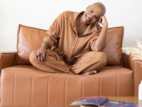 rista Éle Fernandes (mulher preta) sentada de pernas cruzadas em um sofá marrom, ela sorri para a câmera e apoia a cabeça em um dos braços. Exemplares do livro "Dinare" espalhados por uma mesa rendonda baixa e em cima do carpete claro. 