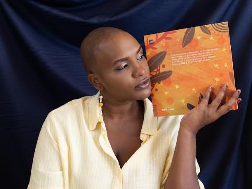 Artista Éle Fernandes (mulher preta) segurando próximo ao rosto um exemplar do livro "Dianari". Ela veste uma blusa amarelo claro de botão, foto com fundo azul escuro.