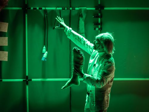 Homem segurando perna artificial humana e apontando para o alto com a outra mão. Veste roupas claras com manga longa e está sob luz verde.