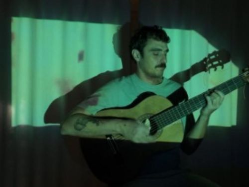 Artista Lucas Caram sentado tocando violão. Iluminação da foto direcionada ao artista.