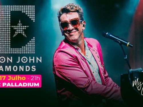 Artista Mizinha Carvalho. Homem sorridente, usando óculos chamativos e blazer rosa, toca piano e canta ao microfone sob luzes de palco.