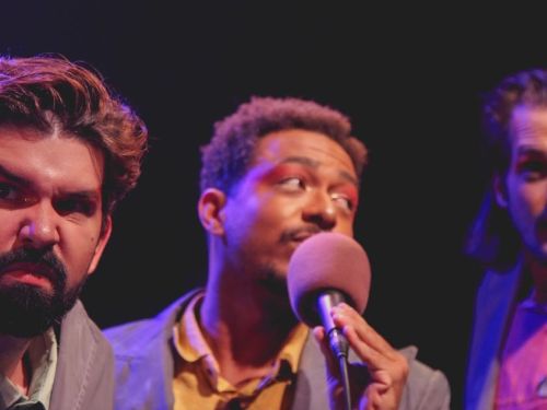 Três atores no palco usando terno e com expressões faciais de seriedade.