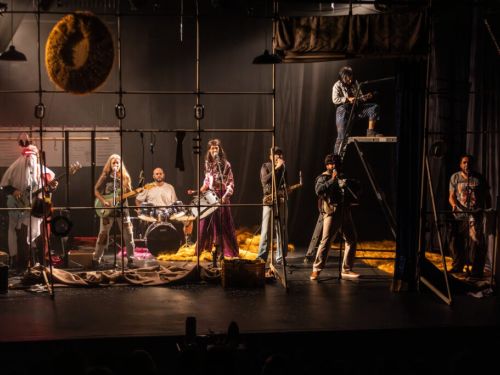 Grupo de teatro cantando em cima do palco. Todos tocam algum instrumento musical e vestem roupas características dos personagens . Palco com iluminação escura e andaimes na frente dos artistas..