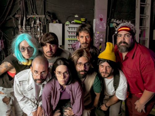 Grupo de teatro posando para foto com semblante sério. Há seis homens e duas mulheres, todos vestem roupas coloridas e características de personagens. Fundo da imagem com equipamentos de som e fiações.