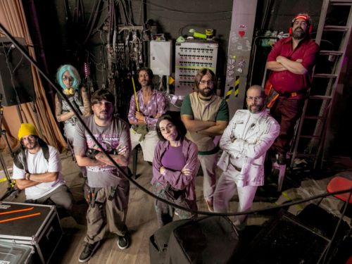 Grupo de teatro posando para foto com semblante sério e os braços cruzados ao peito. Há seis homens e duas mulheres, todos vestem roupas coloridas e características de personagens. Fundo da imagem com equipamentos de som e fiações.