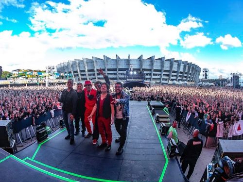 Fotografia em ângulo aberto registrada do palco, mostrando os integrantes da banda Barão Vermelho reunidos à frente de uma multidão imensa de fãs em um festival ao ar livre. Ao fundo, destaca-se a arquitetura imponente do Estádio do Mineirão sob um céu azul com nuvens, capturando a energia do evento com o público de braços levantados.
