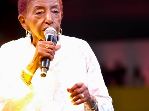 Fotografia vertical da cantora Leci Brandão, uma mulher negra de cabelos curtos avermelhados, capturada em um momento de performance. Ela segura um microfone próximo à boca com a mão direita, enquanto a mão esquerda faz um gesto suave. Veste uma túnica branca com bordados delicados, brincos de argola dourados e diversas pulseiras e anéis. A iluminação de palco incide sobre ela, criando um contraste com o fundo escuro e desfocado.