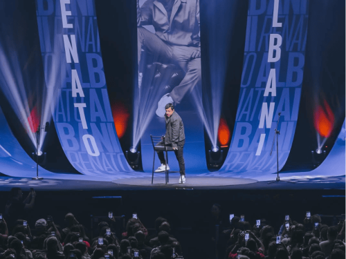  O Gemini disse Fotografia de um show de stand-up do humorista Renato Albani. No palco iluminado por luzes azuis e brancas, ele aparece em pé ao lado de uma banqueta alta. Ao fundo, três grandes painéis verticais exibem seu nome e uma foto sua em preto e branco. À frente do palco, uma plateia lotada é vista de costas, com dezenas de celulares levantados registrando a performance.