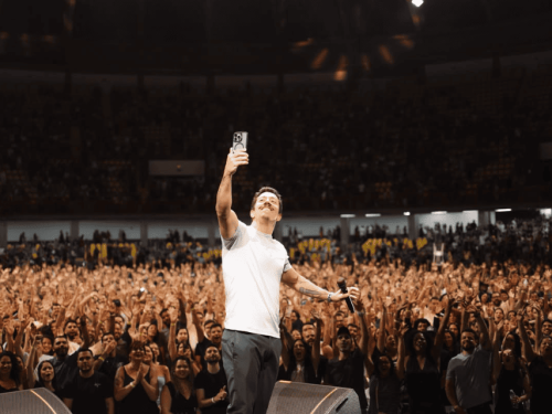 Fotografia de **Renato Albani** no palco, vista de frente para uma plateia imensa e lotada. O humorista, vestindo camiseta branca e calça cinza, segura um microfone em uma das mãos enquanto ergue o celular com a outra para tirar uma **selfie com o público**. Ao fundo, milhares de pessoas aparecem com os braços levantados em um ambiente de arena, criando uma atmosfera de celebração e sucesso.