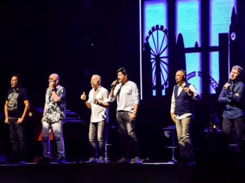 Fotografia do grupo **Roupa Nova** em uma performance ao vivo no palco. Os seis músicos estão perfilados, cantando em microfones sob iluminação cênica. Ao fundo, um telão brilha em tons de azul com a silhueta de uma paisagem urbana, incluindo uma roda-gigante. O ambiente é escuro, destacando os artistas em ação.