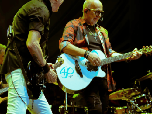  Dois músicos da banda Roupa Nova tocam juntos no palco. À esquerda, um guitarrista de camisa preta e calça branca; à direita, outro integrante toca um violão branco com o logotipo "40 Anos". A cena é capturada em um ângulo dinâmico, com instrumentos de bateria visíveis ao fundo sob iluminação de show.
