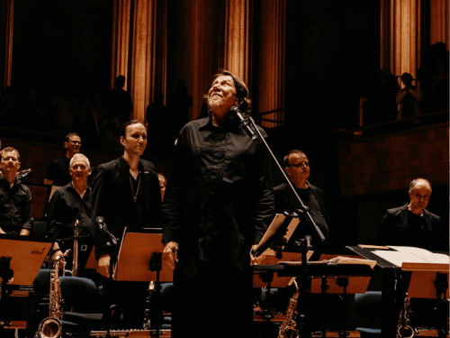 A imagem mostra o cantor Lenine no centro de um palco, olhando para cima com expressão de gratidão ou inspiração. Ele está acompanhado por uma orquestra ou banda de sopros, com músicos posicionados ao fundo e estantes de partitura à frente. O ambiente tem uma iluminação quente e amarelada, destacando grandes colunas clássicas ao fundo, o que sugere uma apresentação em um teatro ou sala de concertos imponente. Todos vestem roupas escuras, reforçando o tom solene e artístico da cena.